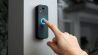 A person presses the illuminated button on a modern smart video doorbell mounted on a wall.
